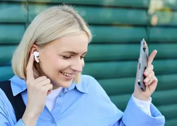 Blonde Frau mit iPhone in einer Hand, während sie mit dem Zeigefinger der anderen Hand auf ihr Ohr mit einem Apple AirPods Pro 2 zeigt; grüne Wand im Hintergrund