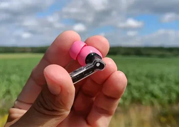Rezensent hält einen rosa Nothing-Ear-(3a)-Ohrhörer in der Hand, mit Ackerland im Hintergrund
