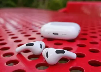 Apple AirPods Pro 3 auf einem perforierten roten Stahltisch liegend, mit dem Ladecase dahinter