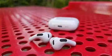 Apple AirPods Pro 3 auf einem perforierten roten Stahltisch liegend, mit dem Ladecase dahinter