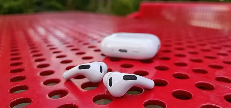 Apple AirPods Pro 3 auf einem perforierten roten Stahltisch liegend, mit dem Ladecase dahinter